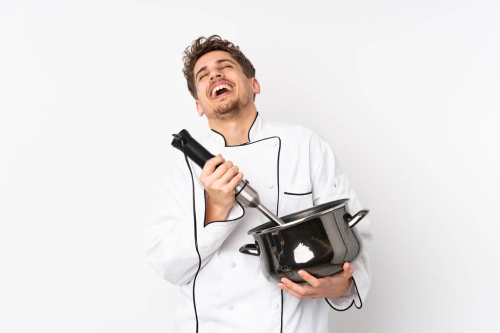 about me 1 A man in his 30s wearing a beige apron gestures toward an open kitchen cabinet filled with old mixers, with an air fryer visible on the counter behind him.